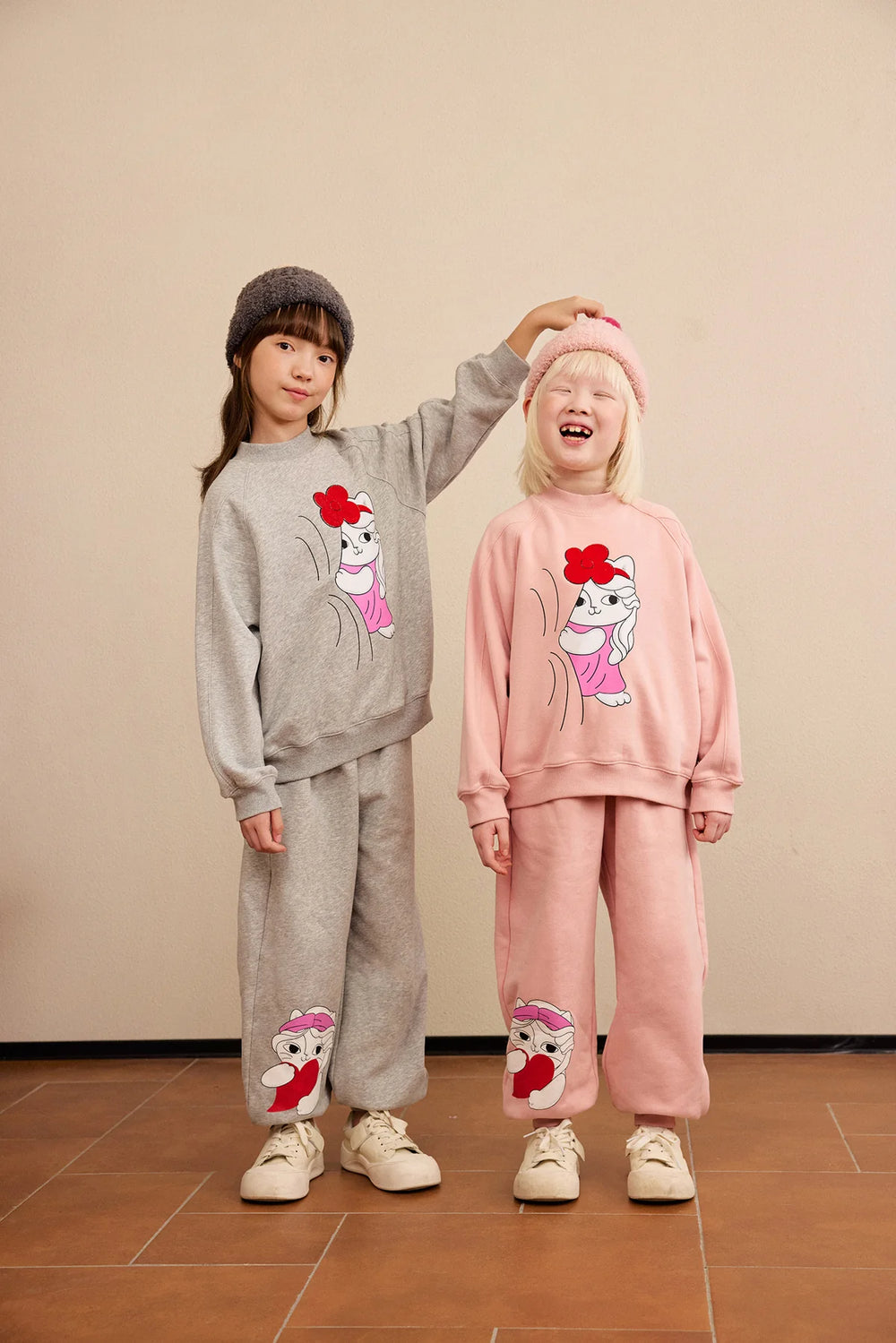 Two children in the Hide-and-Seek Cat sweatshirt and joggers, one in grey and one in pink.
