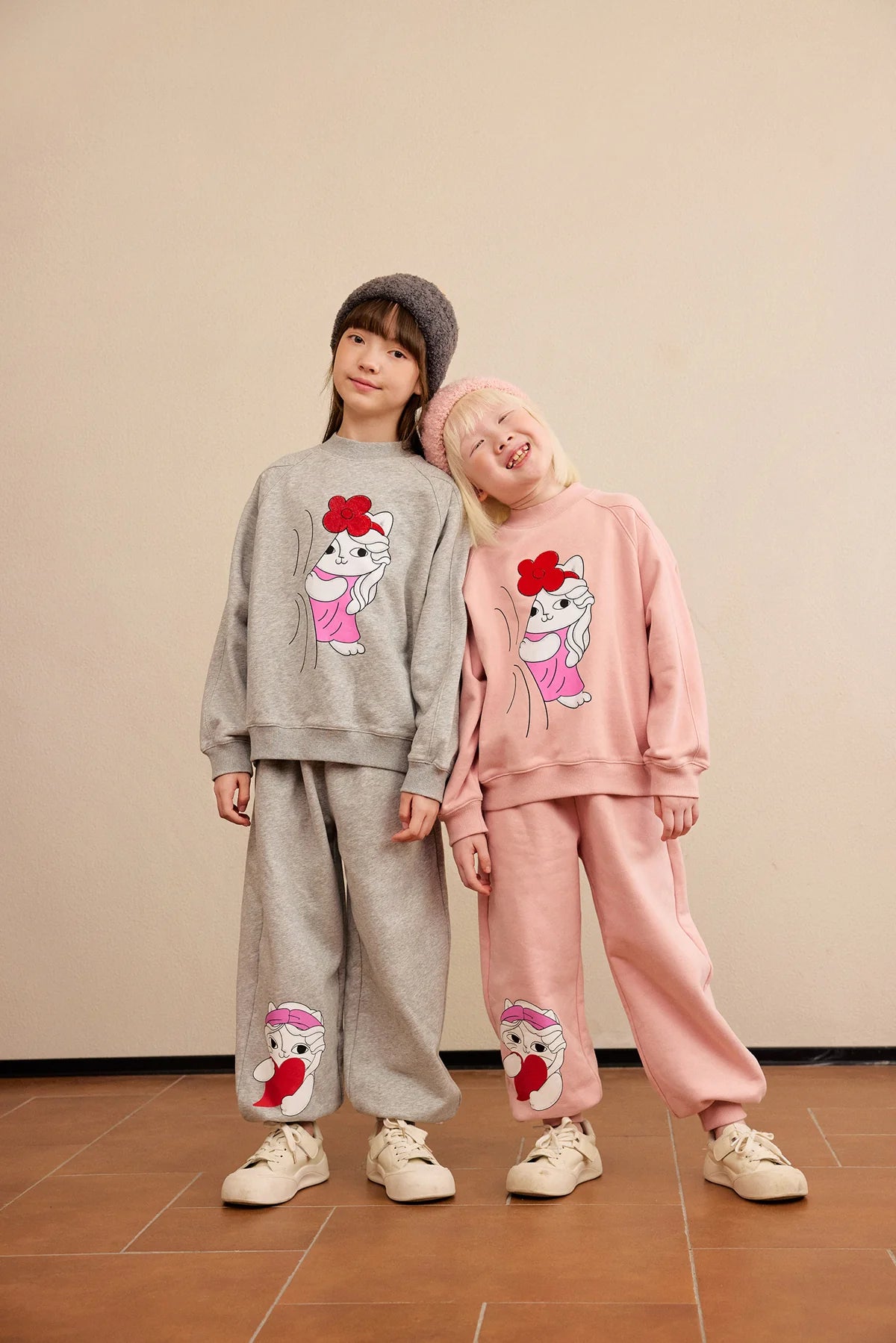 Two children leaning together in grey and pink Hide-and-Seek Cat sweatshirts and joggers.