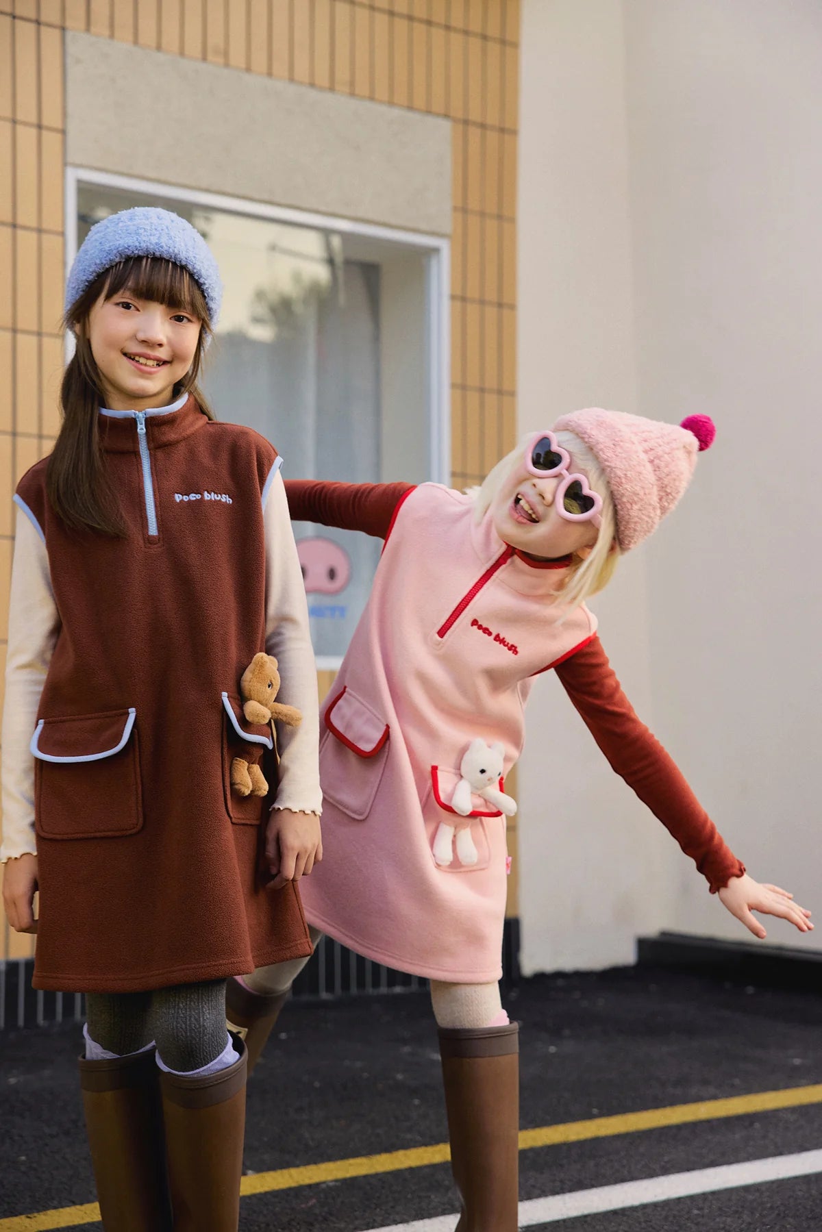 Two children in the Animal-Pocket Fleece Pinafore, in caramel brown and dusty pink.
