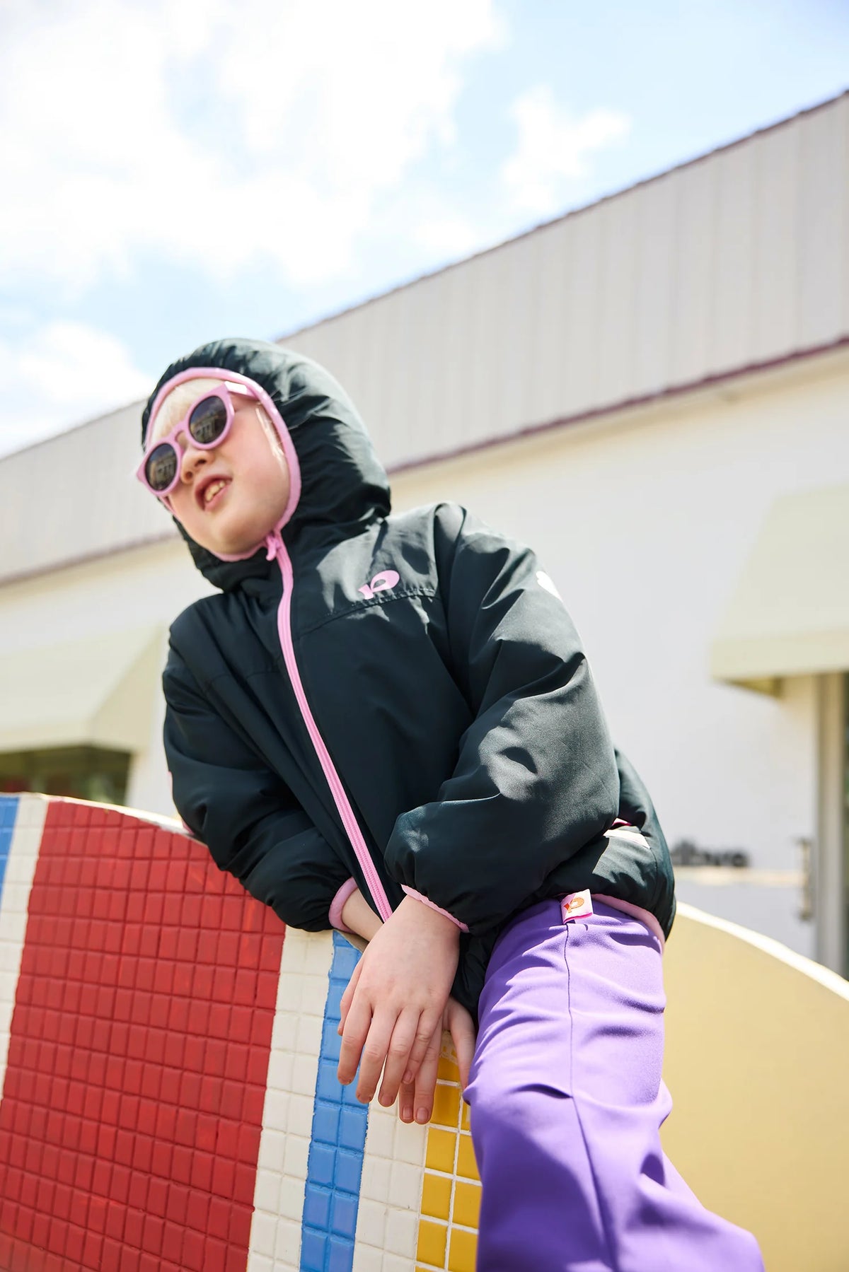 A child in the black reversible hooded jacket sits on a colourful bench, seen from the front.